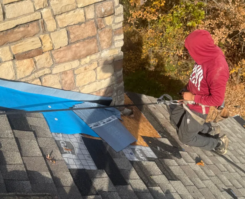 Worker repairing a shingle roof beside a stone chimney.